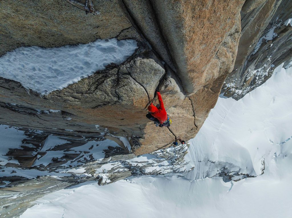 Climber in red jacket navigating a steep rock crack with snow-covered slopes visible below.