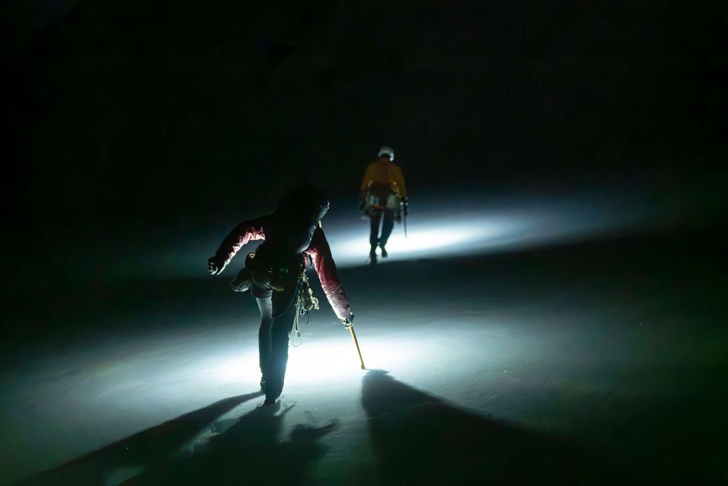 Two mountaineers hiking in darkness guided by bright headlamps across a snow-covered glacier at night.