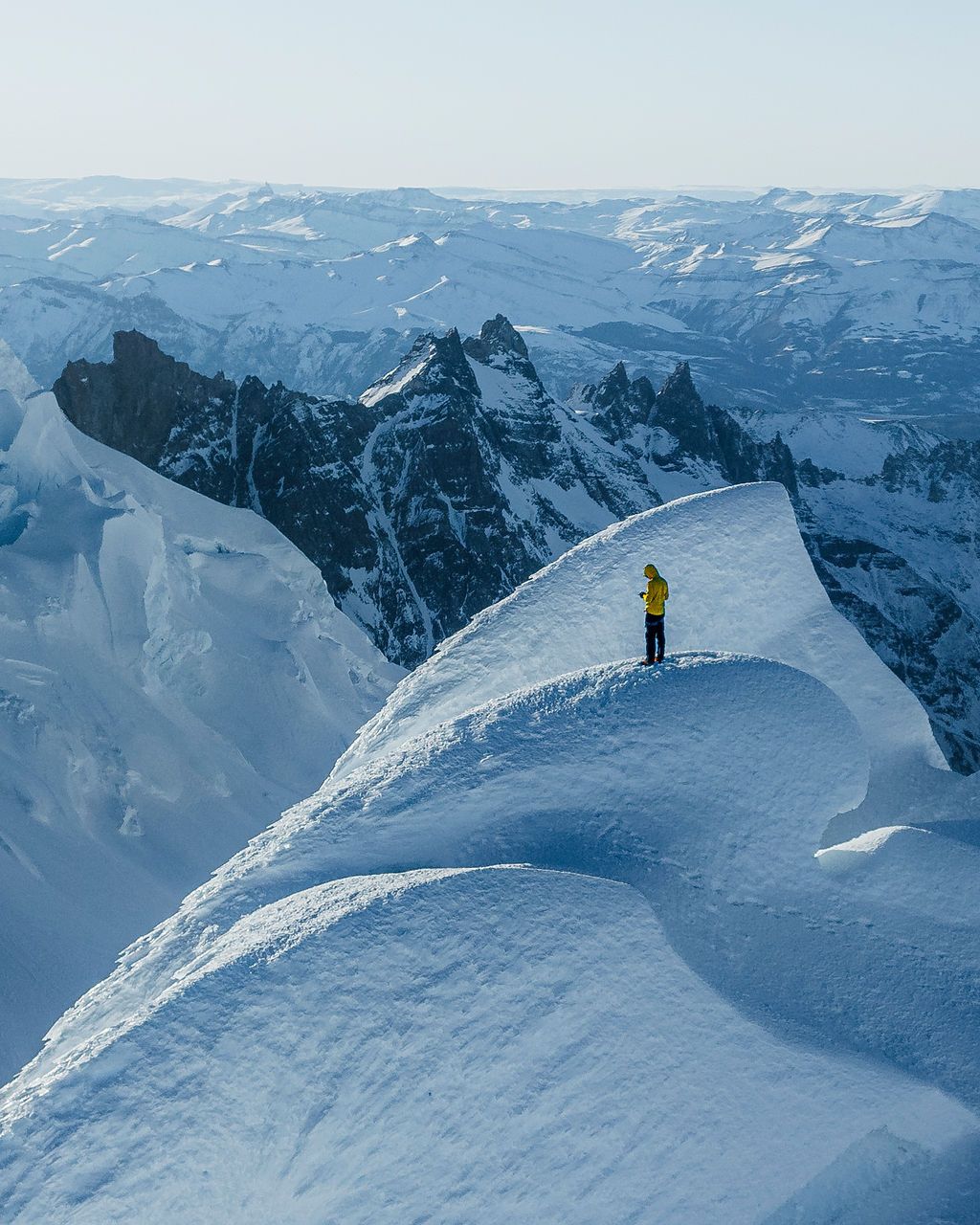 A lone mountaineer in a yellow jacket stands atop a snow ridge surrounded by vast snowy mountain ranges.
