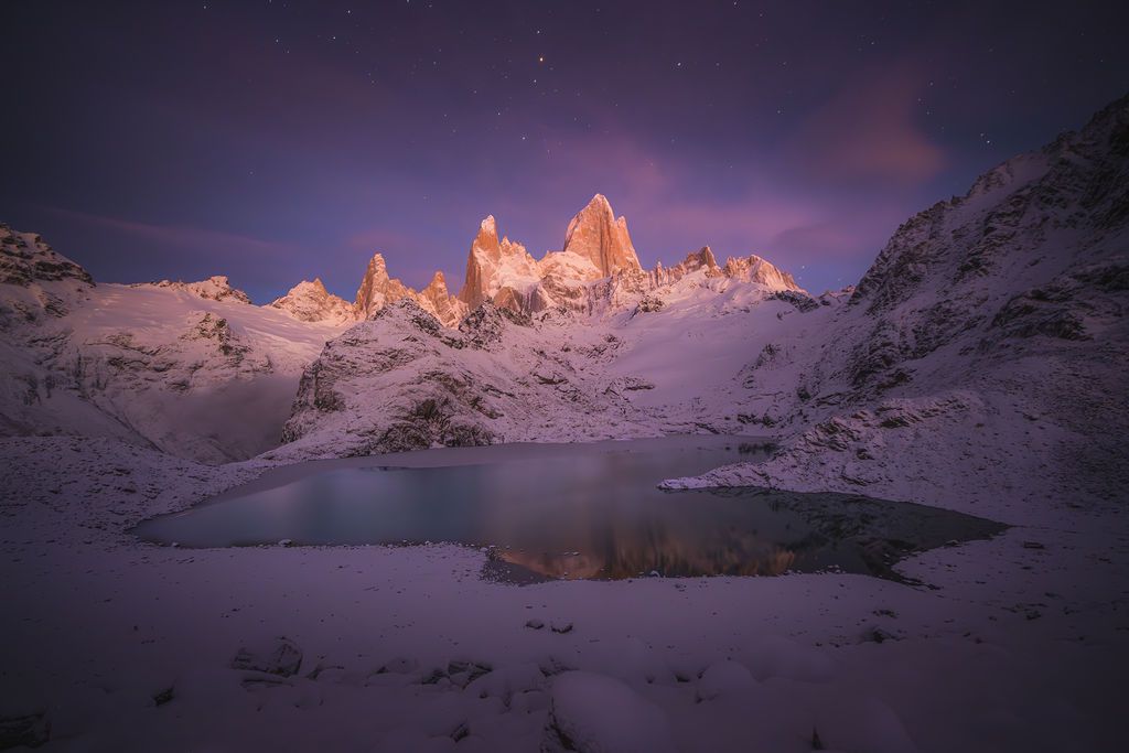 Purple twilight sky over snow-covered Fitz Roy mountain reflected in a frozen alpine lake.