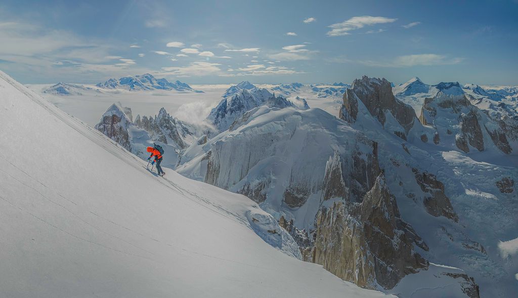 Skier descends steep snow slope with dramatic Patagonian mountain range and clouds in the background.