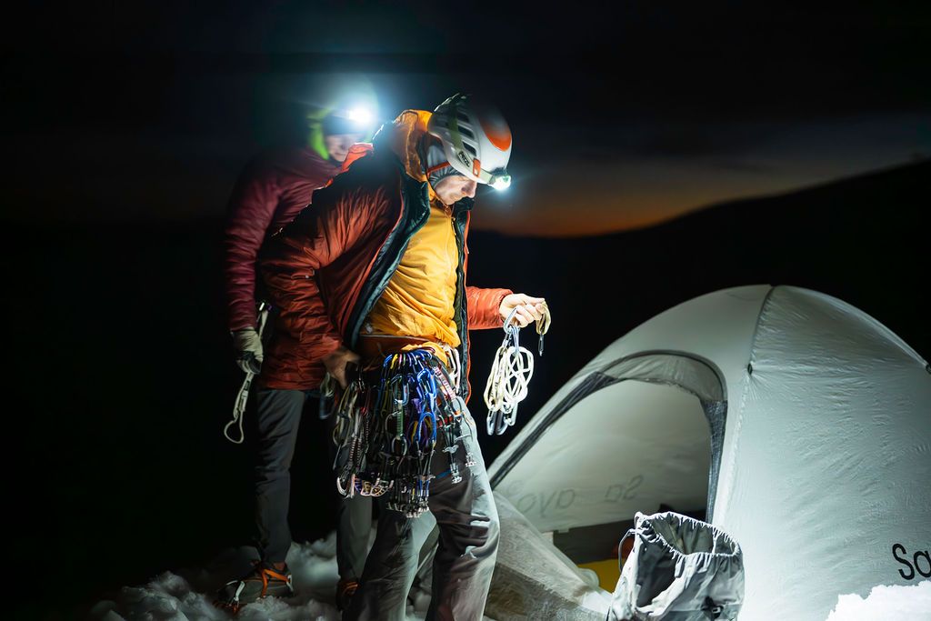 Two mountaineers in helmets preparing climbing gear outside a tent in darkness at high altitude.