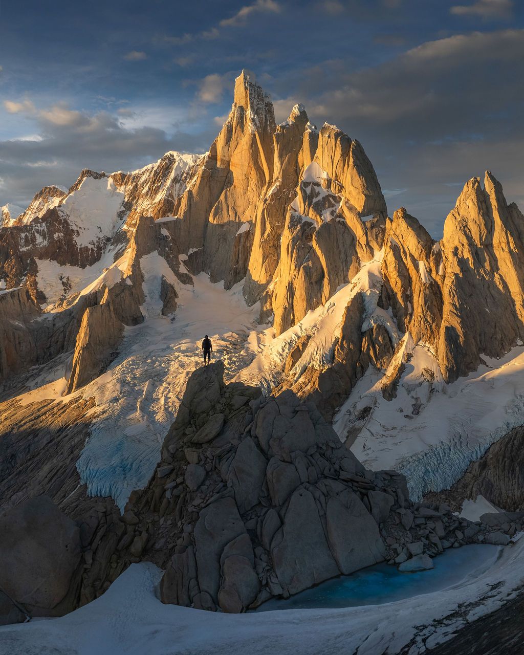 Lone hiker stands on rocky outcrop before towering Mount Fitz Roy at golden sunset in Patagonia.