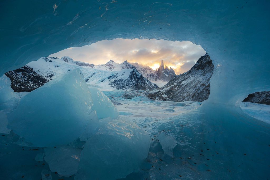 Snow-covered Fitz Roy mountain peaks are dramatically framed through a glacial ice cave at golden hour.