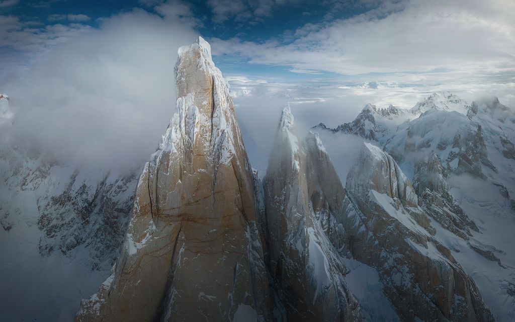 Aerial close-up of snow-dusted granite spires of Cerro Torre emerging through swirling clouds in Patagonia.