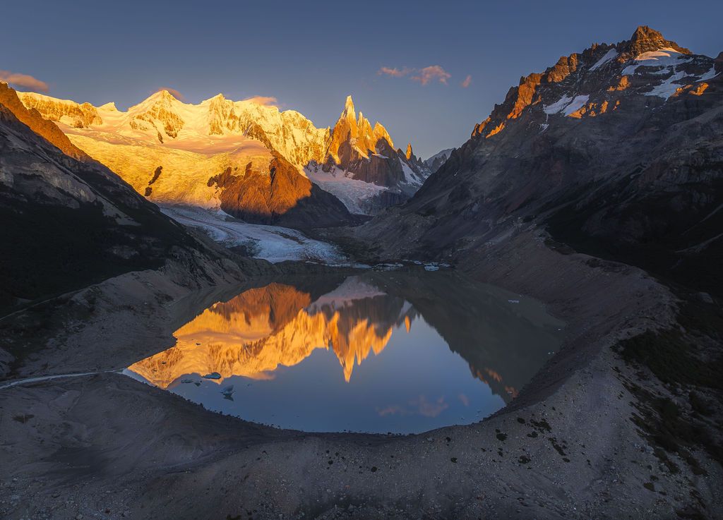 Golden alpenglow reflects perfectly in a calm glacial lake below Cerro Torre peaks at sunrise.