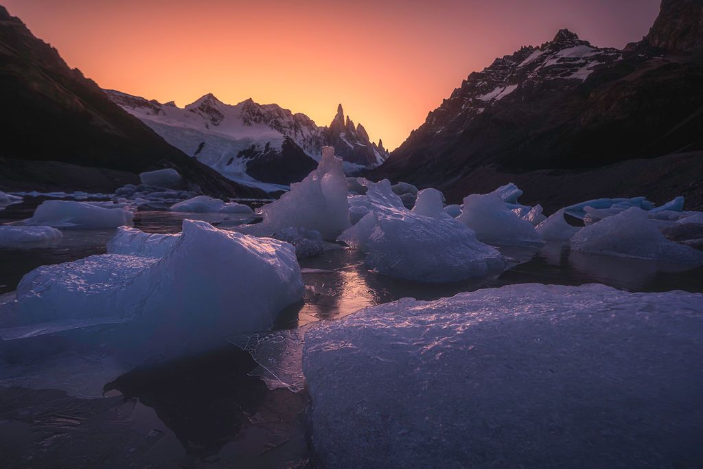 Vibrant orange sunset glows behind jagged mountain peaks surrounded by icebergs in Patagonia.