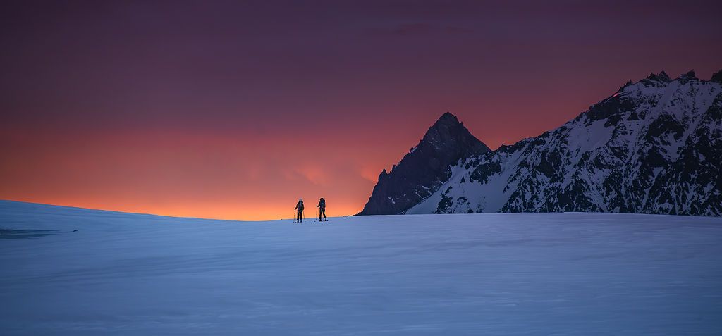 Two silhouetted hikers traverse a snowy ridge at sunset with a dramatic orange and purple sky in Patagonia.