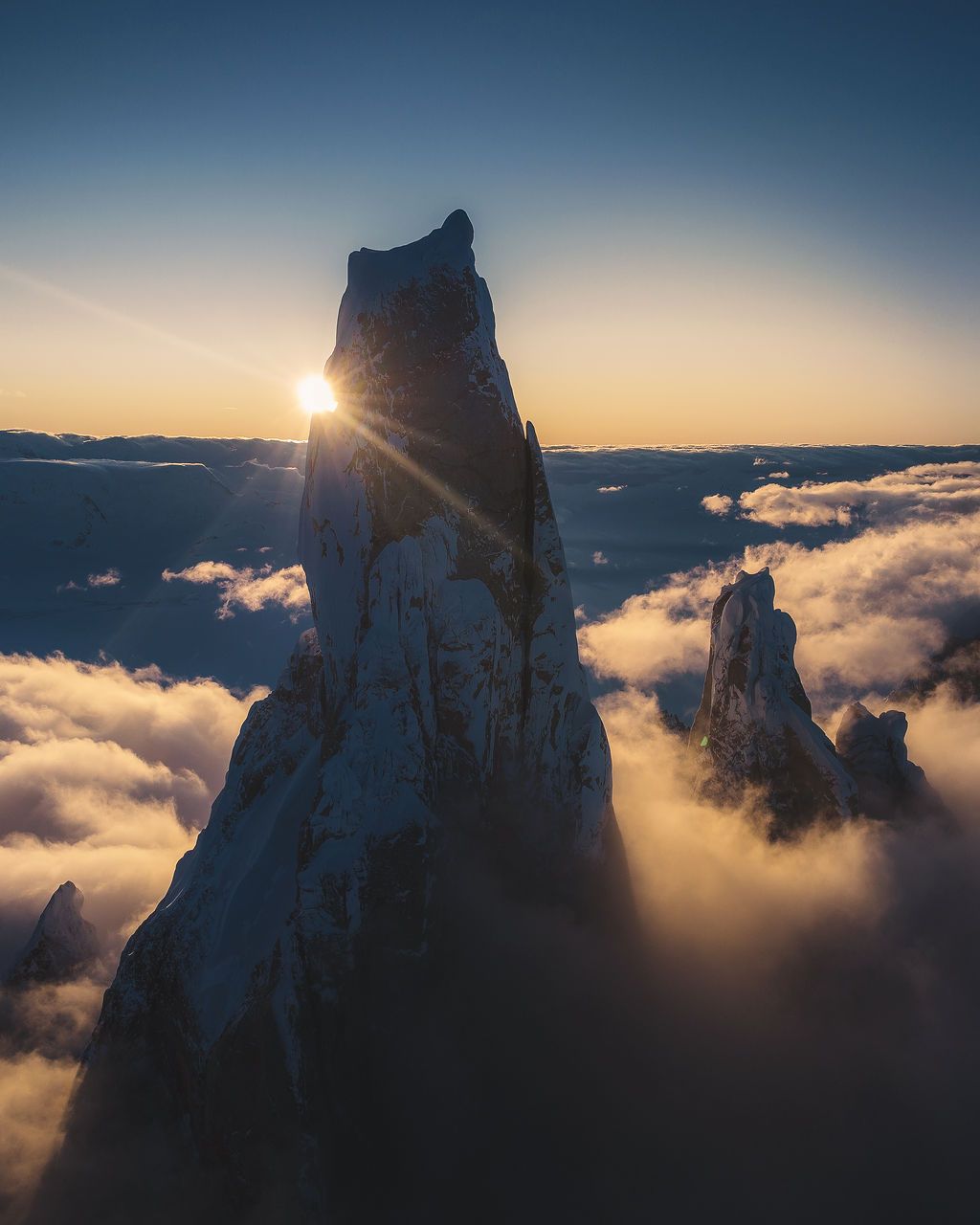 A dramatic snow-capped mountain spire rises above a sea of clouds at golden sunset from an aerial perspective.