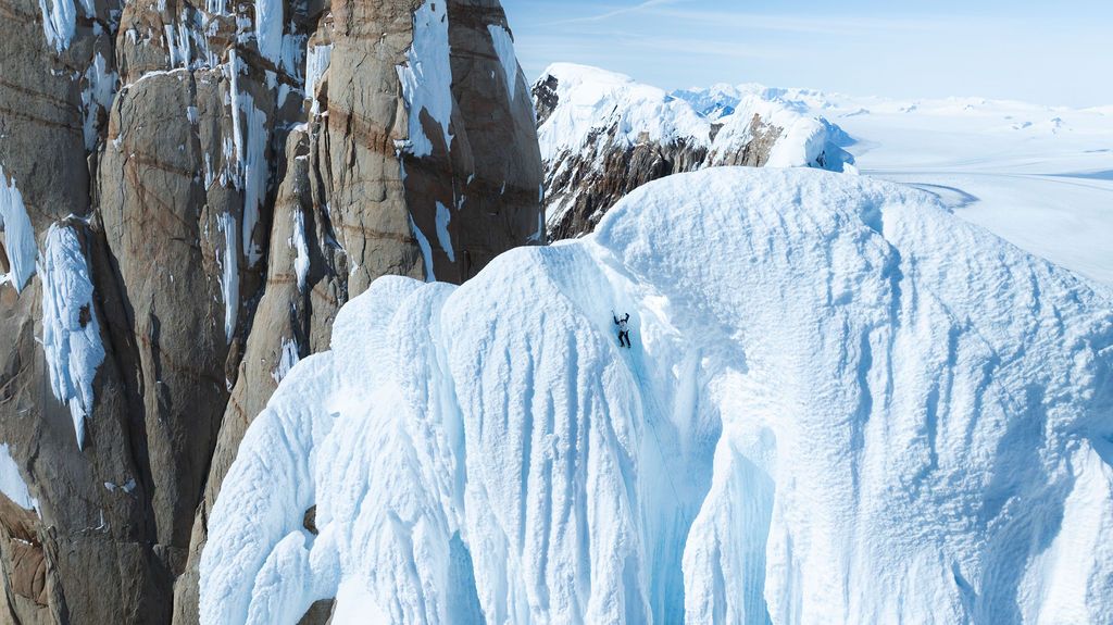 Aerial view of climbers ascending a massive ice wall with snow-covered landscape stretching to horizon.