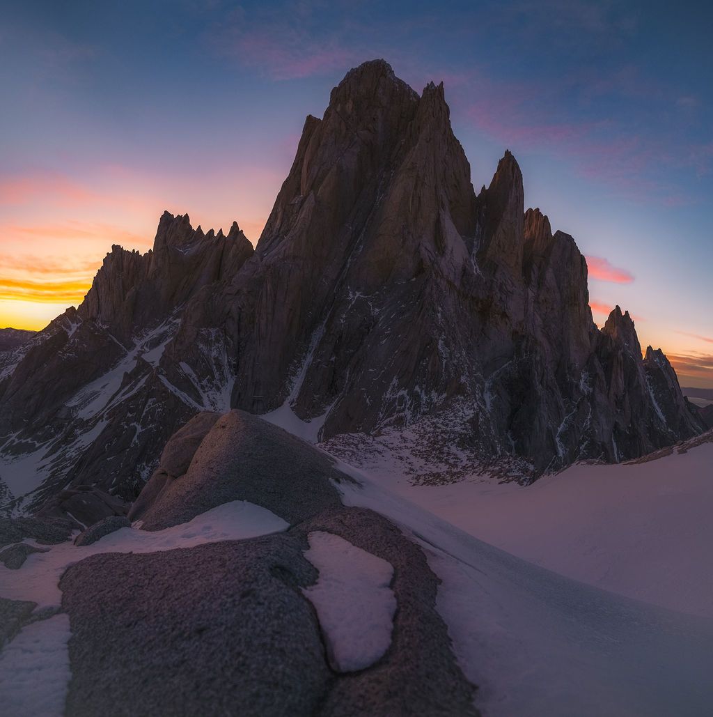 Colorful sunset sky illuminates sharp rocky mountain peaks covered in snow.