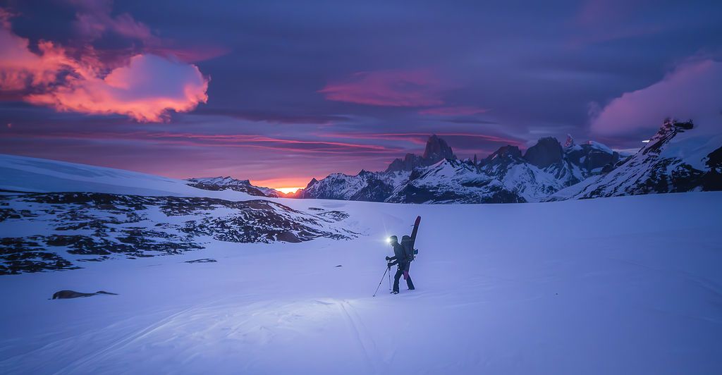 Solo mountaineer with headlamp treks across snowy glacier at dusk beneath vivid purple Patagonian sky.