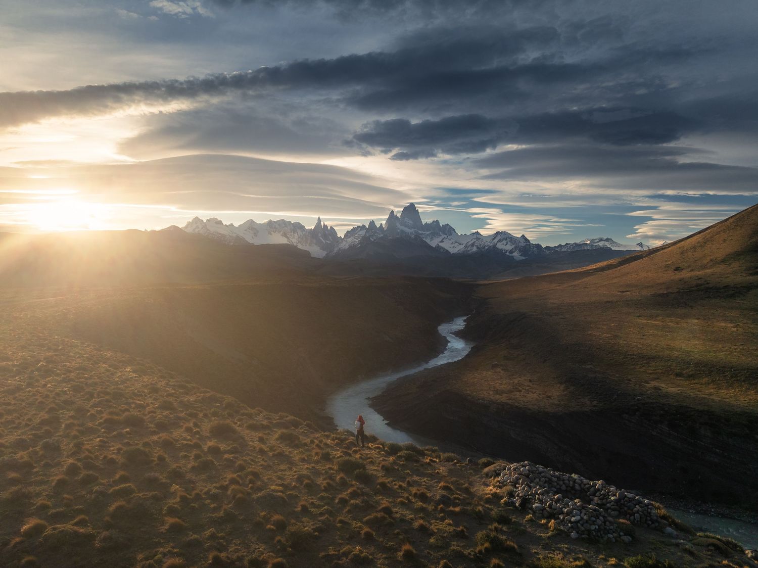 Aerial sunset view of a winding river through golden Patagonian plains with jagged snow-capped peaks under dramatic clouds.