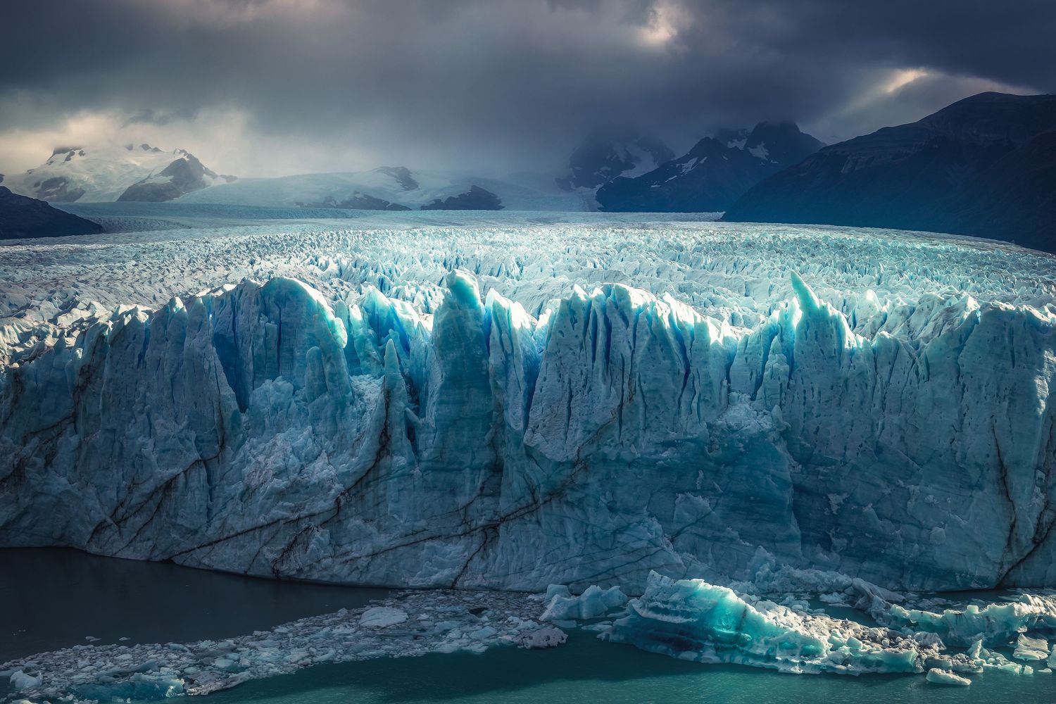 Dramatic blue glacier with jagged ice formations meets dark stormy skies and mountains in Patagonia, Argentina.