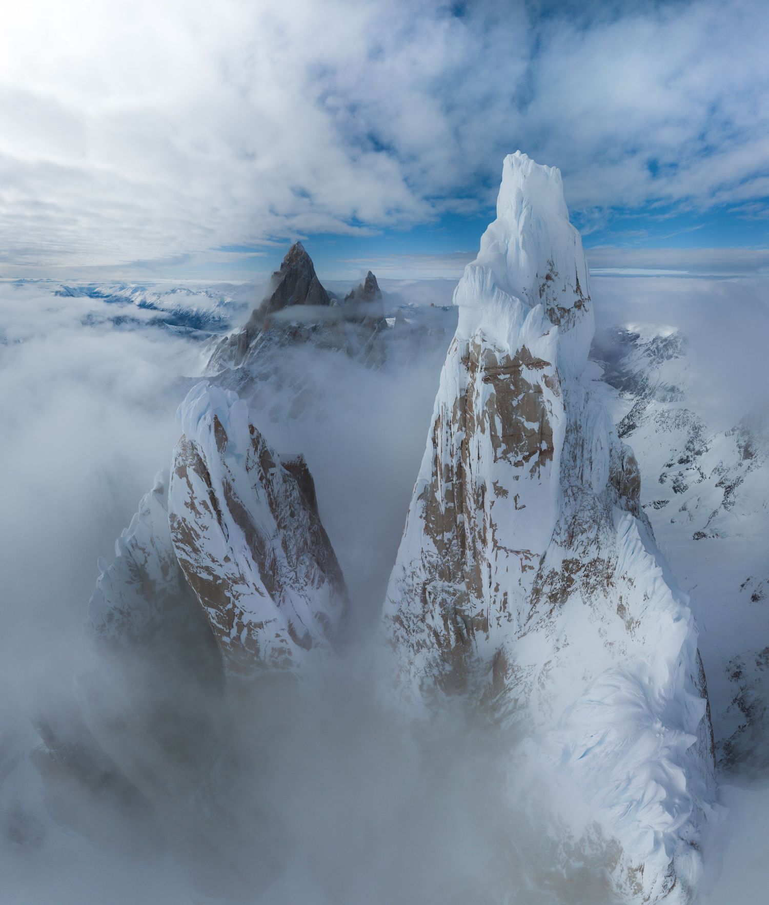Aerial view of snow-covered jagged mountain peaks piercing through clouds in a dramatic alpine landscape.