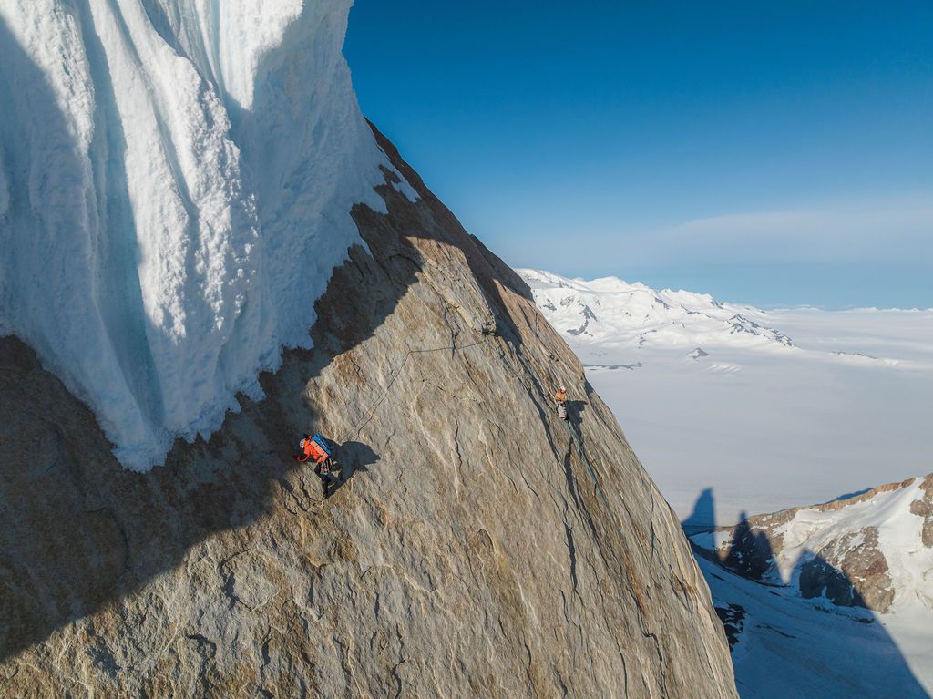 Two climbers traversing a rocky ridge with massive ice formations and vast snowy plains below.