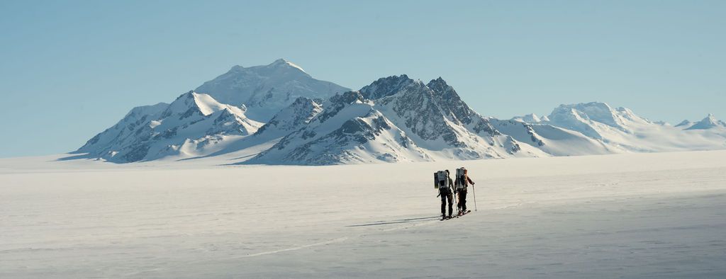 Two hikers trekking across a vast white snow-covered landscape with mountains in the distance.