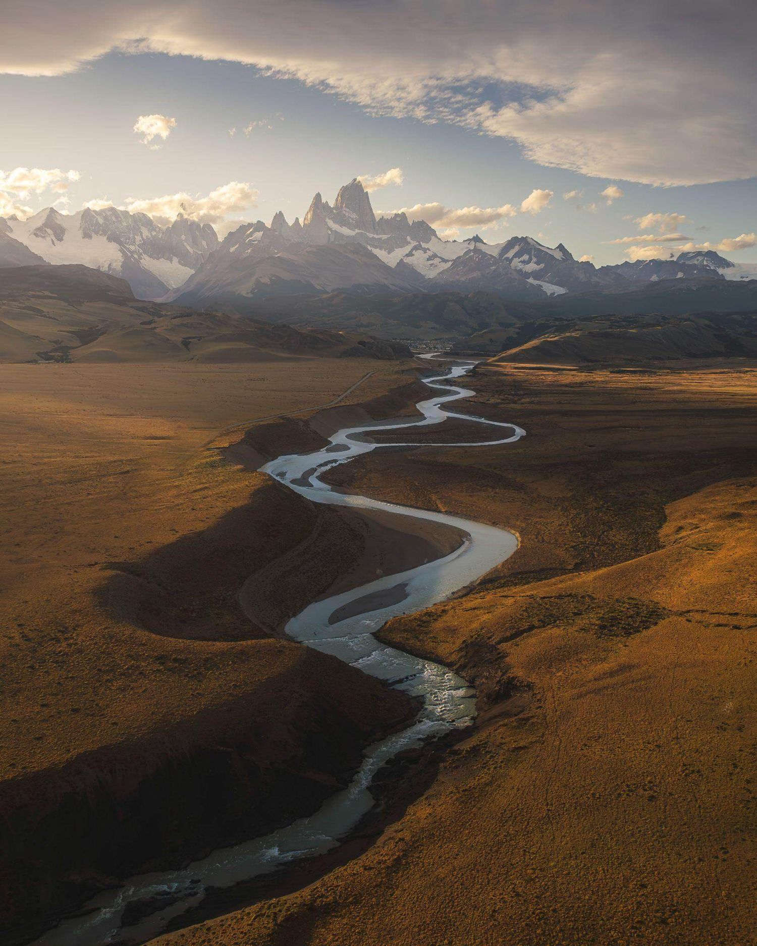 Aerial view of a winding river crossing golden Patagonian steppe with Mount Fitz Roy in the background.