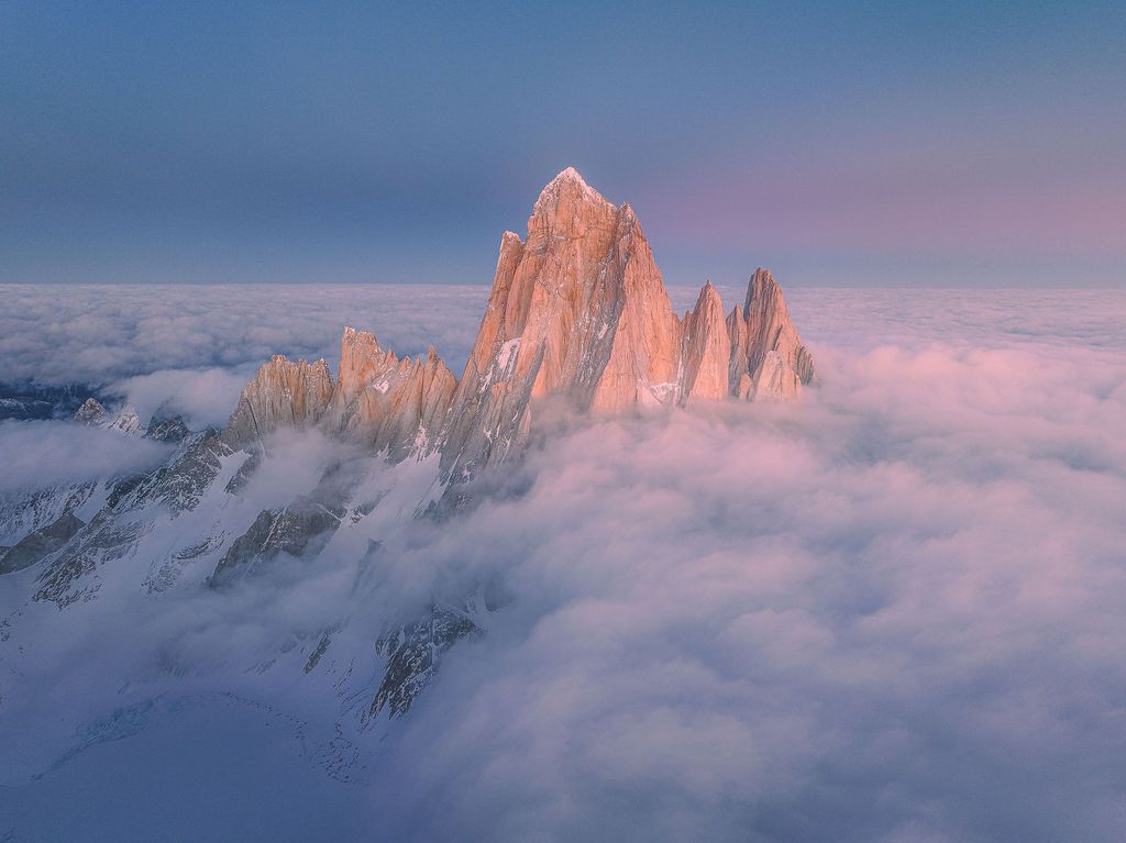 Aerial view of Mount Fitz Roy's sharp granite spires rising above a sea of clouds at pink sunset.