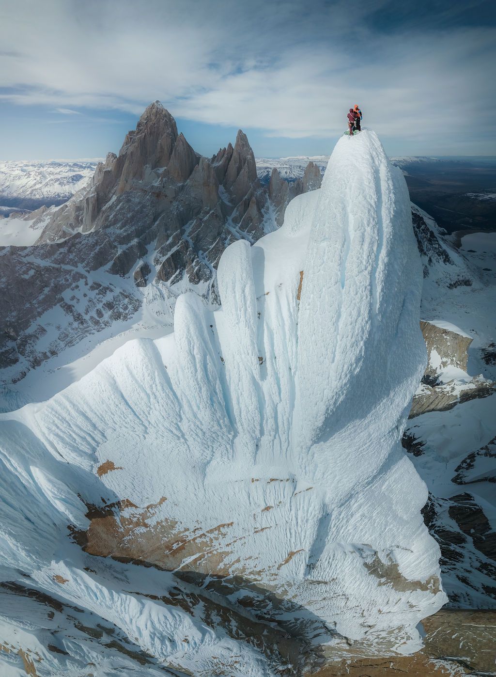 Climbers atop a dramatic snow-covered rock pinnacle with jagged mountain peaks visible in the distance.