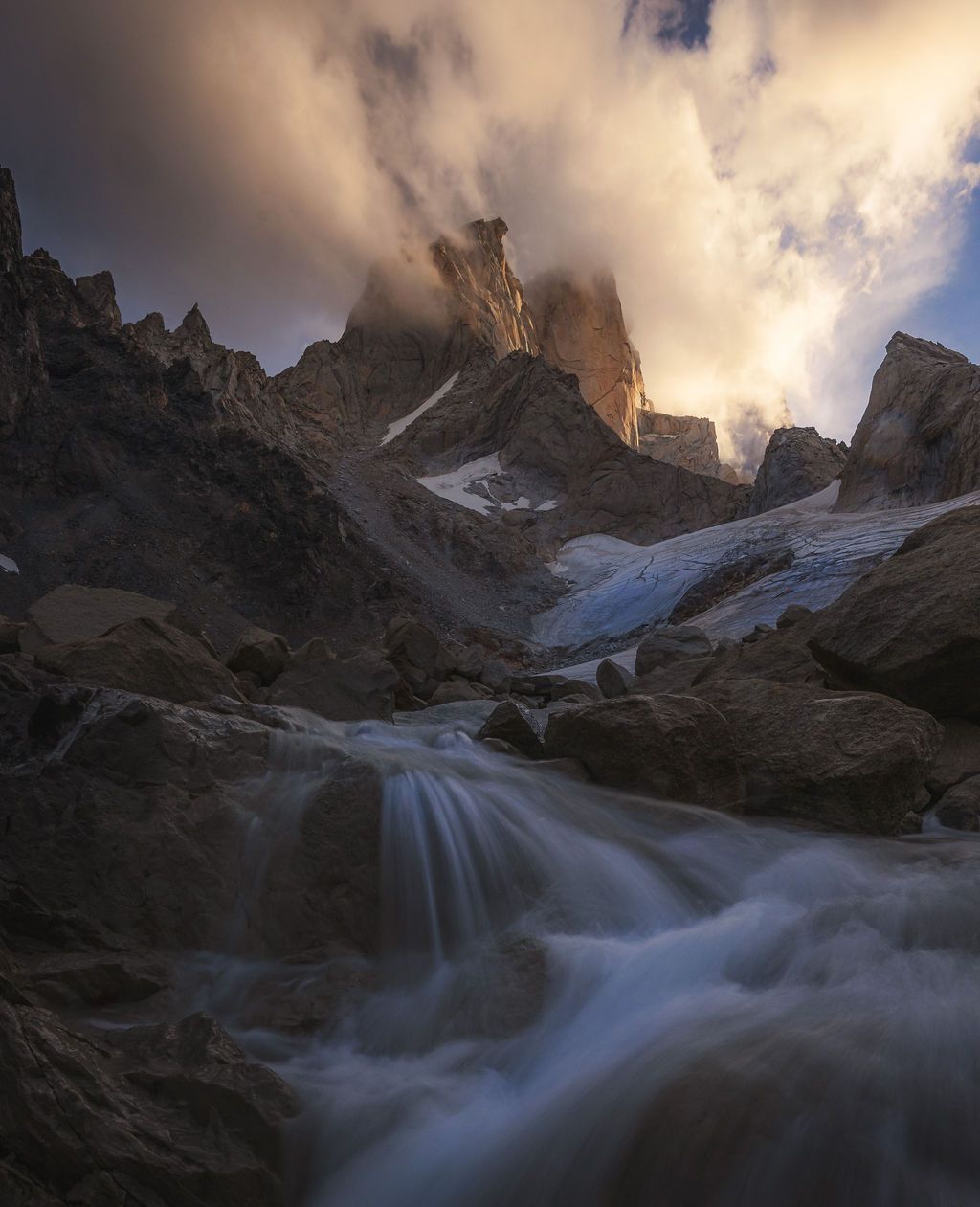 Long-exposure waterfall rushes through rocky terrain beneath dramatic stormy mountain peaks.