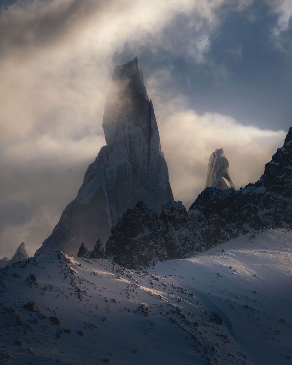 Snow-dusted jagged mountain peaks pierce through dramatic swirling clouds in Patagonia.