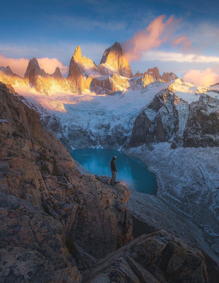 Hiker stands on rocky ledge overlooking turquoise glacial lake at sunrise below Mount Fitz Roy, Patagonia.