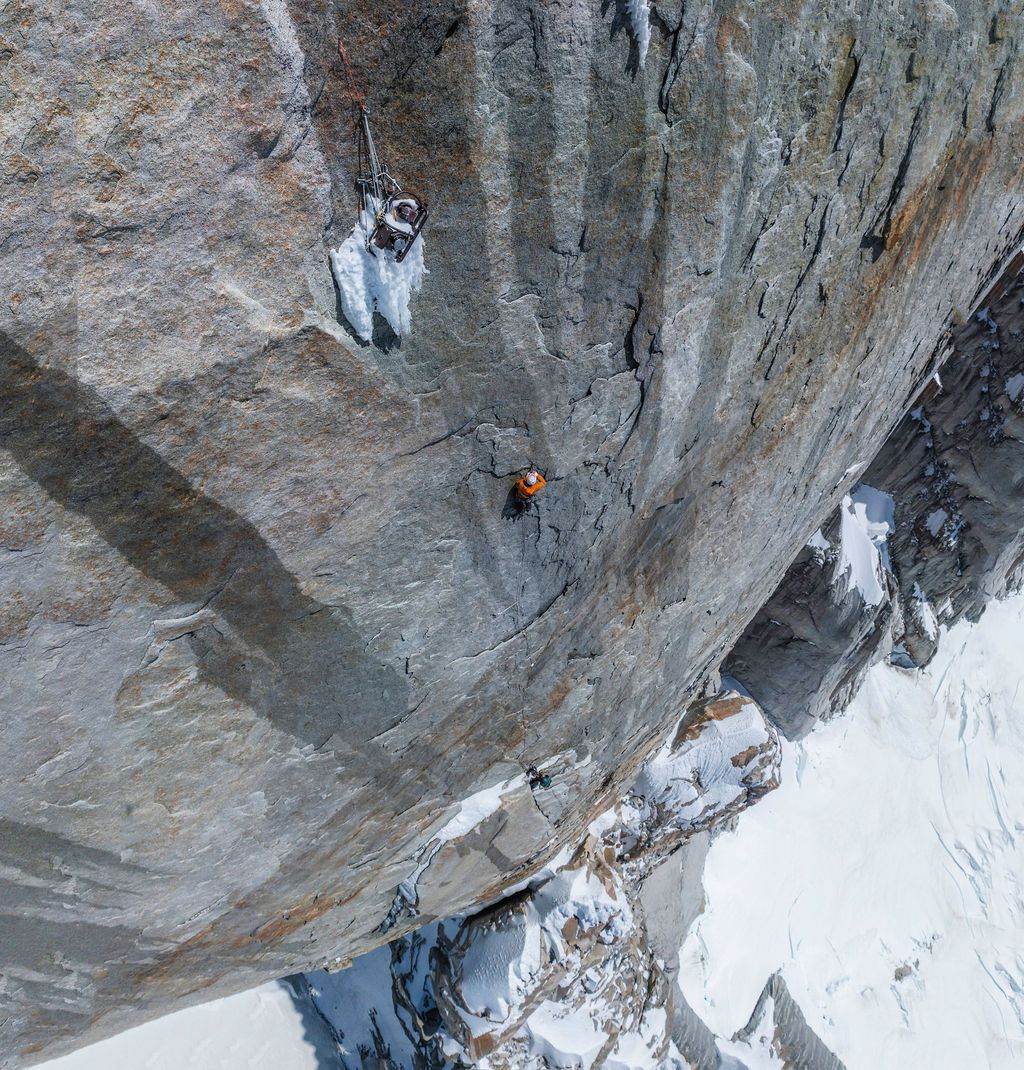 Rock climber in orange gear ascends a steep granite cliff face with icy glacier visible below in Patagonia.
