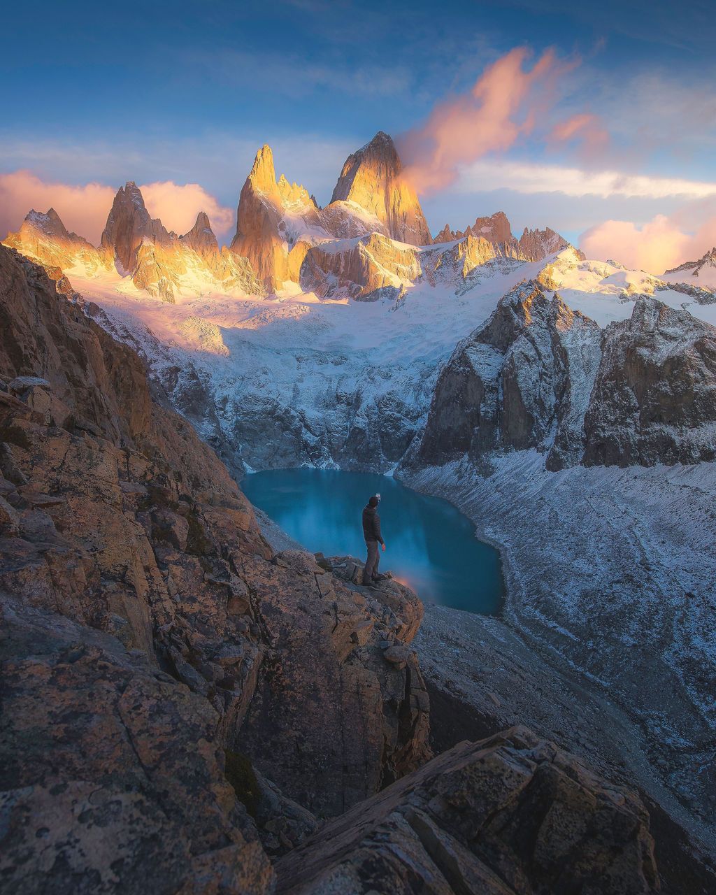 Hiker stands on rocky ledge overlooking glacial lake with snow-capped Fitz Roy peaks glowing at sunrise in Patagonia.
