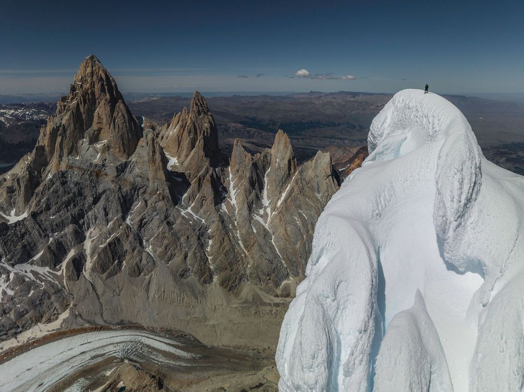 Climber stands atop an ice-covered peak surrounded by jagged granite towers of Patagonia under blue sky.