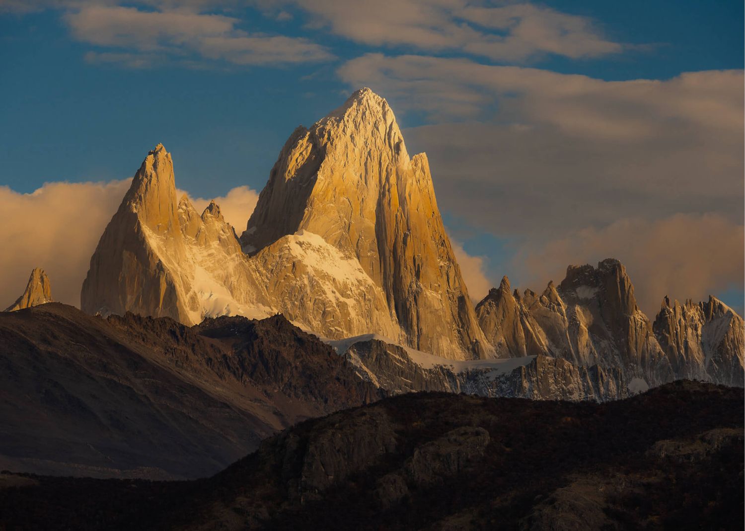 Golden morning light illuminates the iconic granite spire of Mount Fitz Roy rising above clouds in Patagonia.