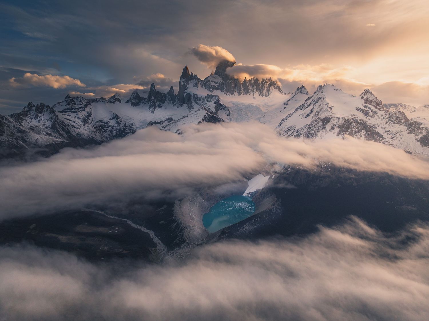 Aerial view of snow-capped Patagonian peaks emerging through dramatic clouds at golden sunset with turquoise glacial lake below.