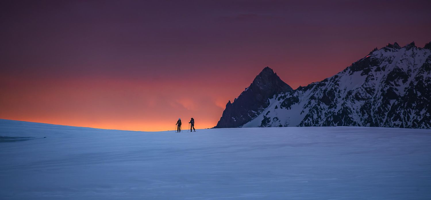 Two silhouetted hikers traverse a vast snowy plateau beneath a vivid purple and orange sunset sky.
