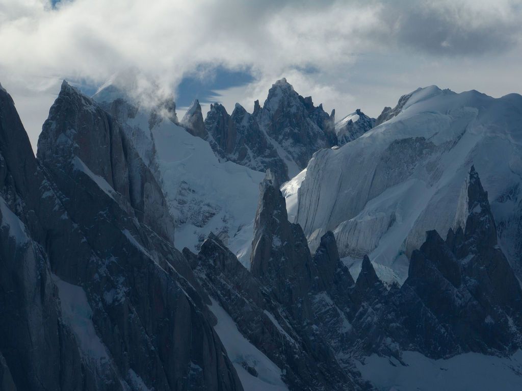Cold blue aerial view of jagged snow-dusted mountain spires shrouded in clouds in Patagonia.