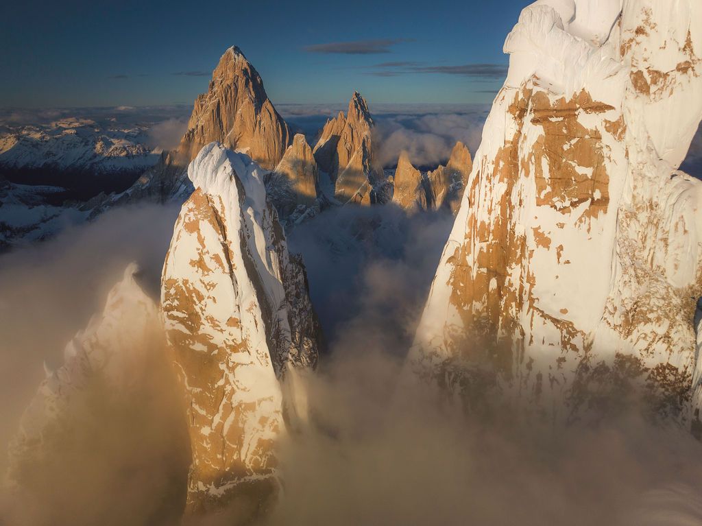 Aerial view of golden sunlit granite spires rising through swirling clouds in the Patagonian Andes.