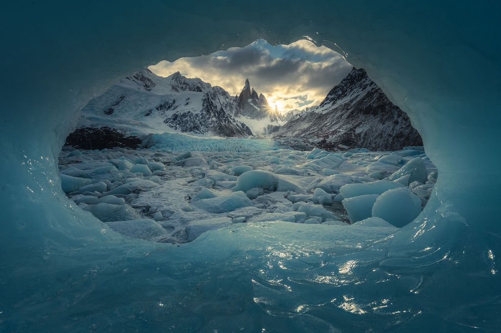 A stunning view of snow-capped Patagonian peaks framed through a natural ice cave opening at sunset.