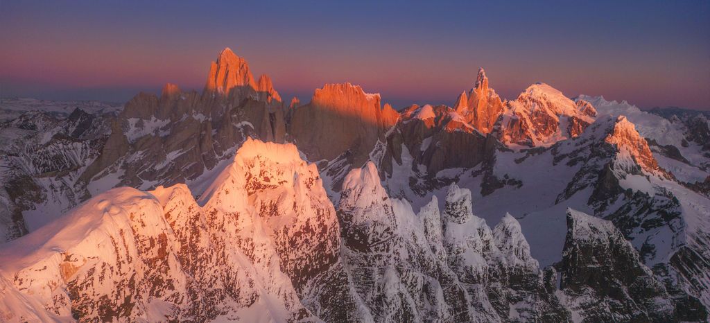 Aerial panoramic view of jagged snow-covered Patagonian peaks glowing orange at sunset above the clouds.