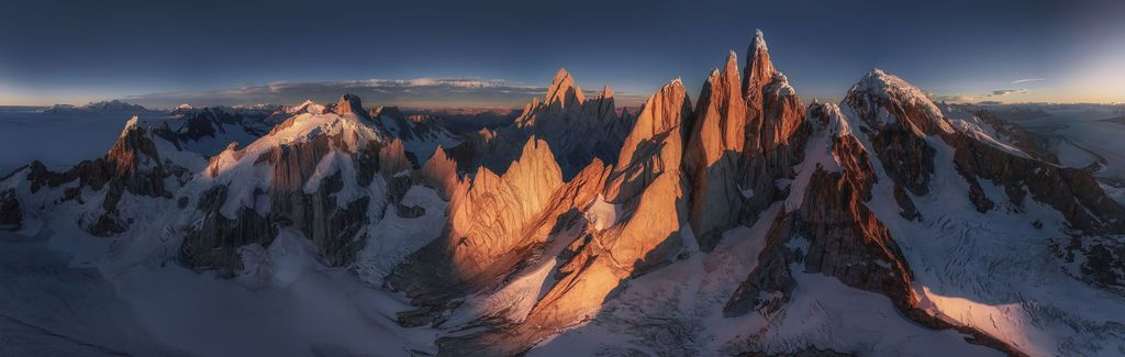 Wide panoramic aerial view of Cerro Torre and Fitz Roy peaks glowing at golden sunrise.