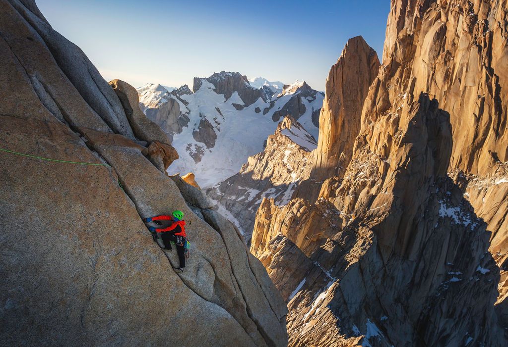 Rock climber scaling a steep granite wall with snow-covered mountain peaks and spires visible in the distance.