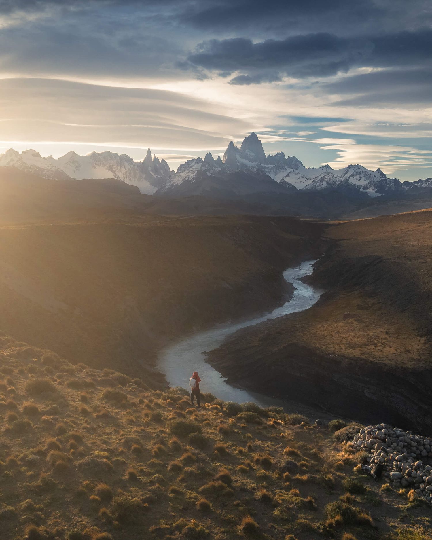 Hiker stands in golden Patagonian valley at sunset with winding river and snow-capped Fitz Roy peaks in background.