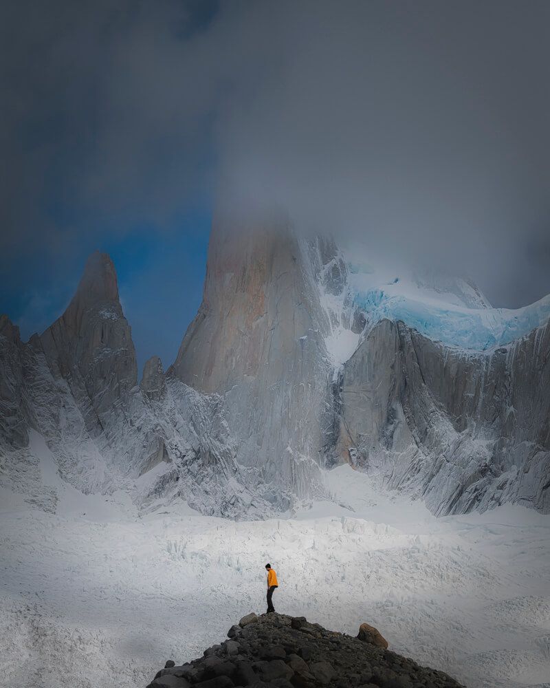 Lone hiker in orange jacket stands before towering snow-covered granite spires of Cerro Torre in stormy Patagonia.