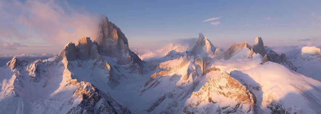 Panoramic aerial view of snow-dusted Patagonian mountain peaks emerging through soft clouds at golden hour.