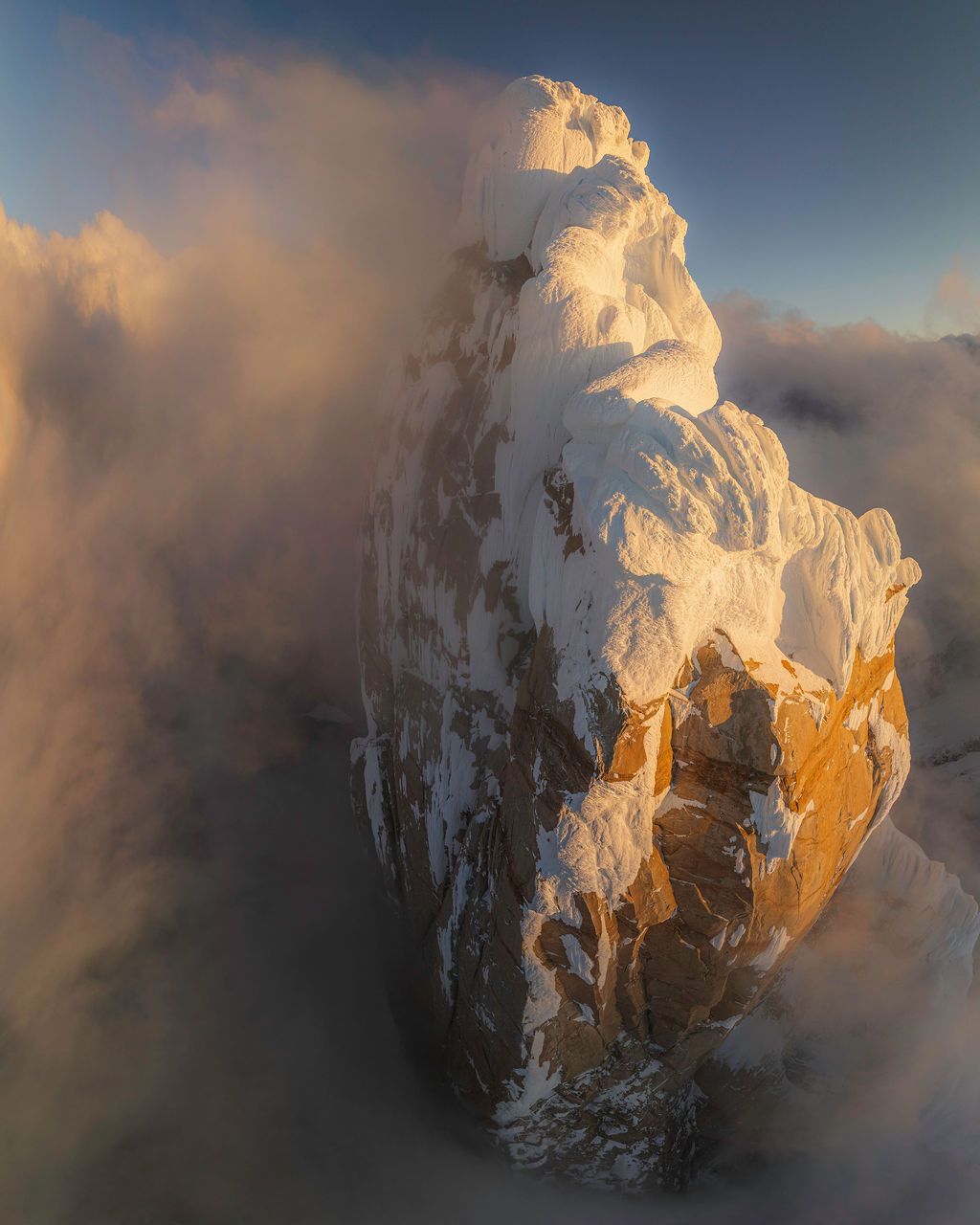 Golden sunrise light illuminates a massive snow-covered mountain peak emerging through clouds.