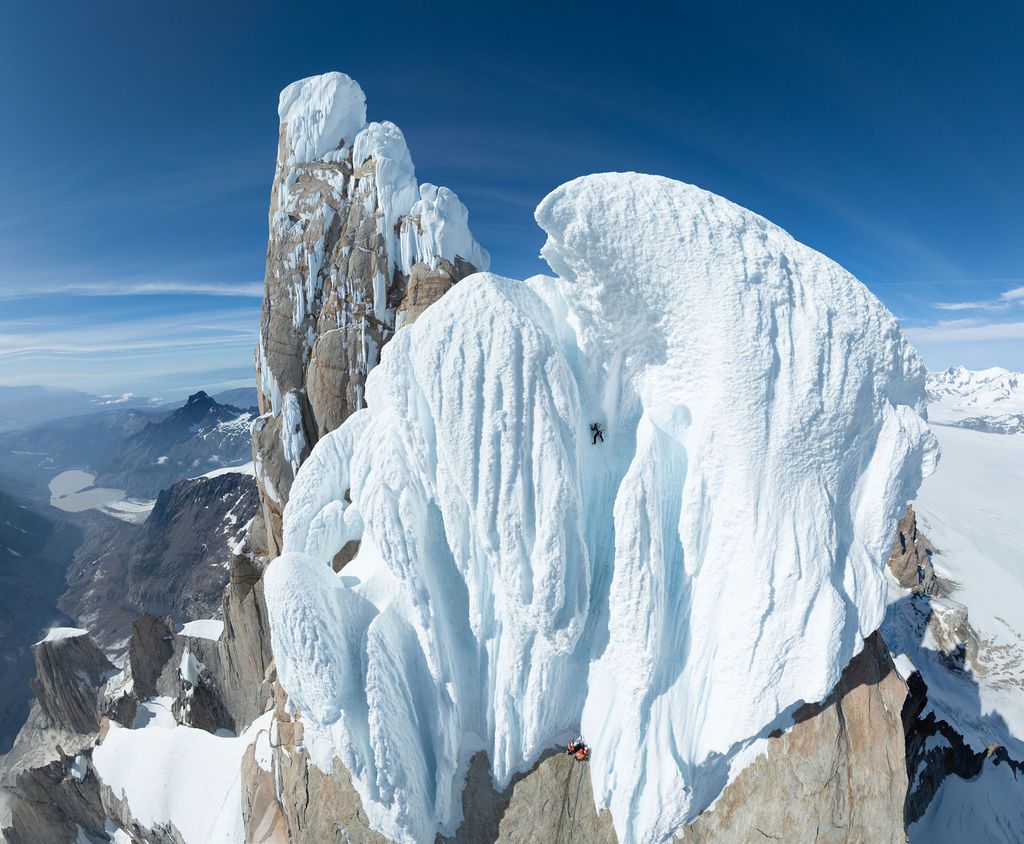 Climber ascending a massive ice mushroom formation on a rocky Patagonian peak under a clear blue sky.