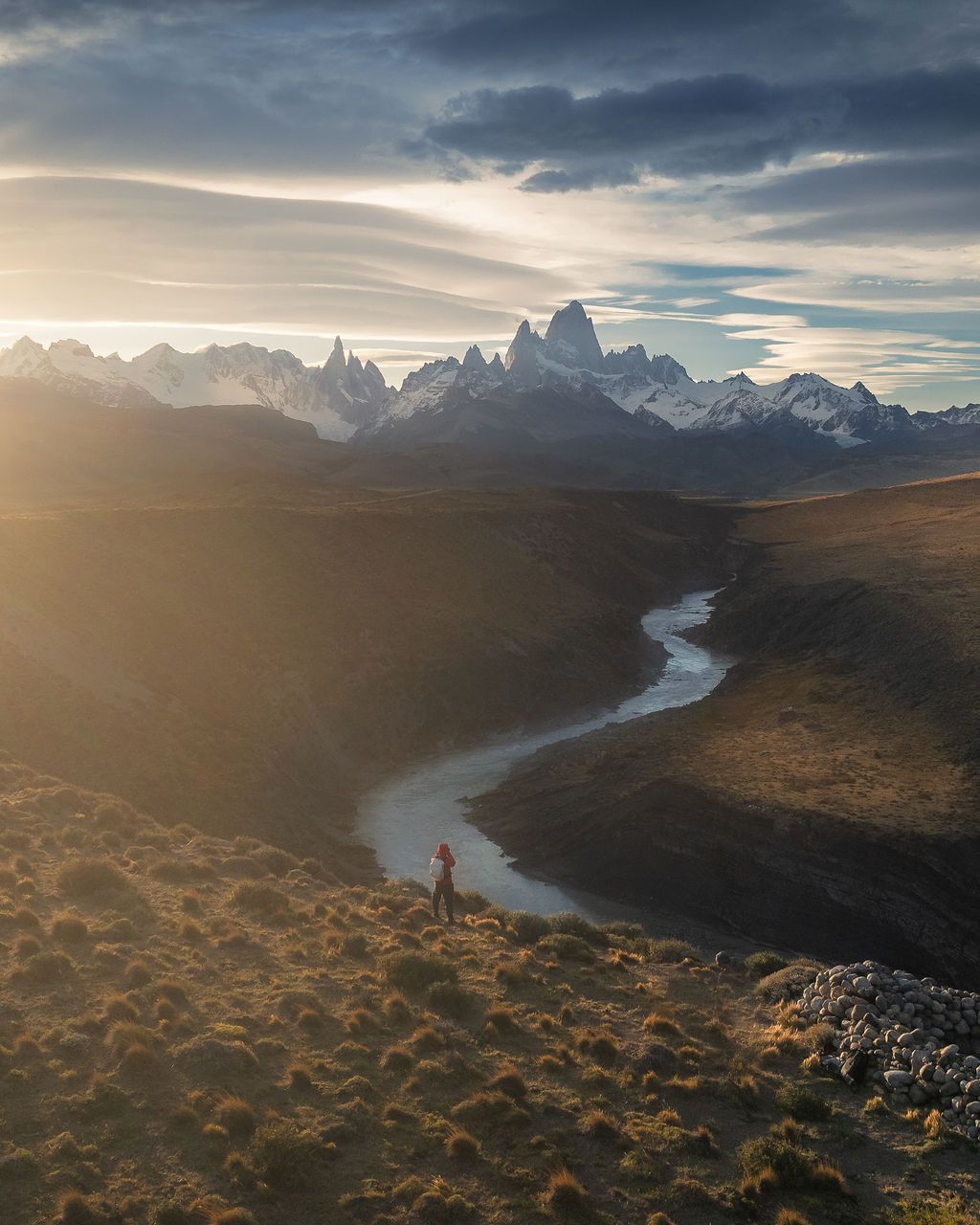 Hiker silhouetted against golden sunset light with winding river leading to Fitz Roy peaks.