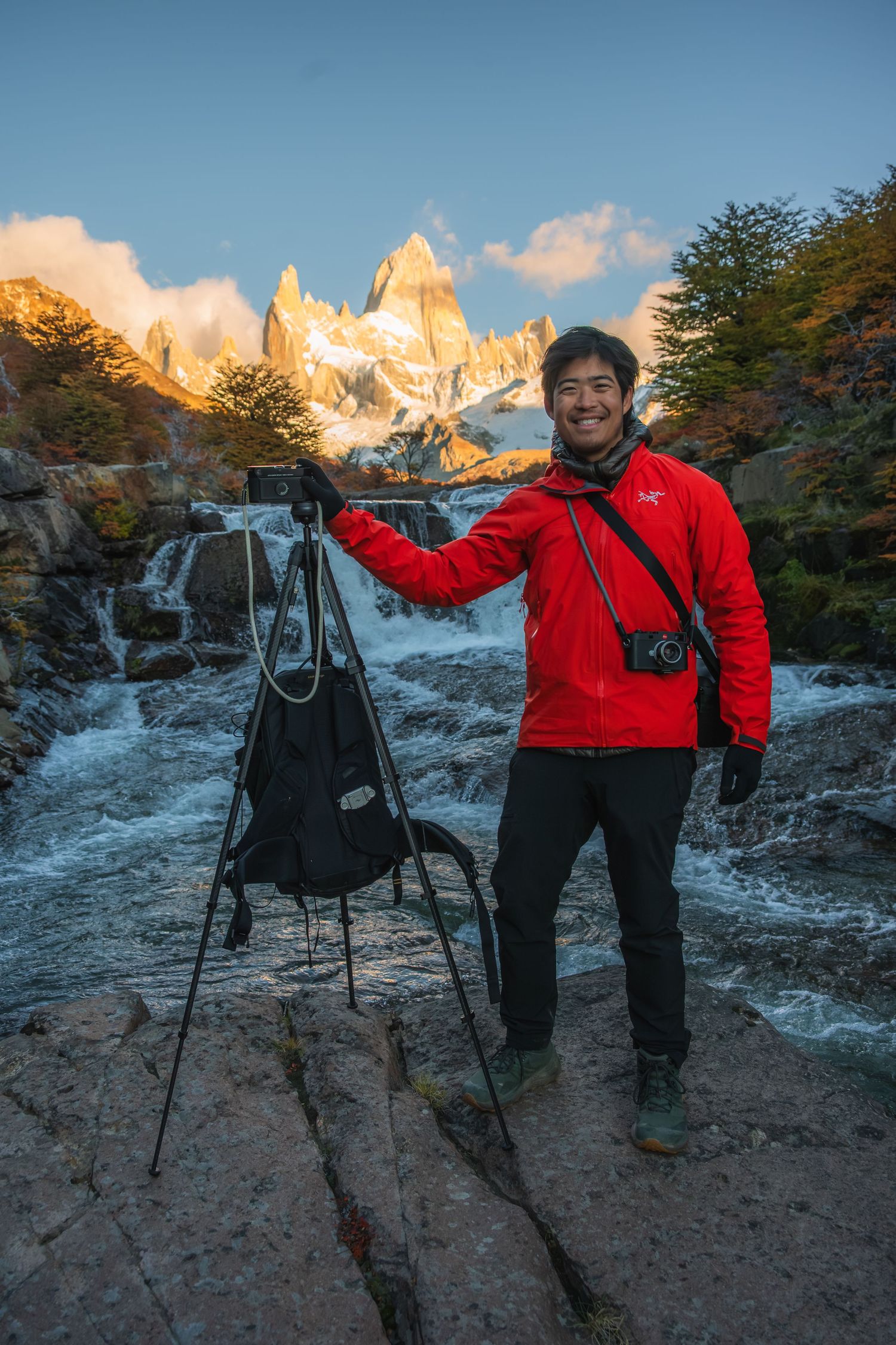 Smiling photographer in red jacket poses with tripod camera gear beside a waterfall with snowy mountain peaks in Patagonia.