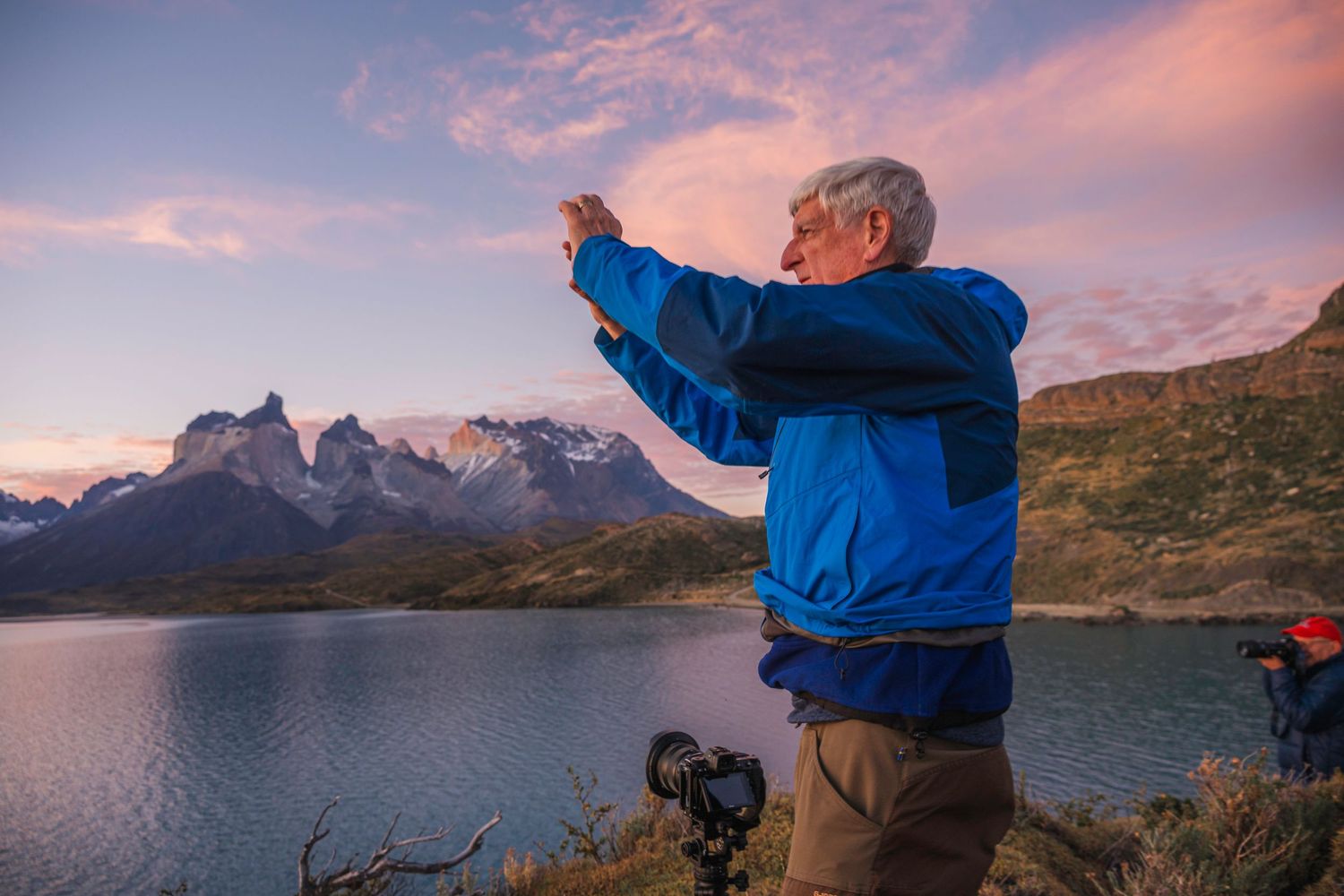 Man in blue jacket photographing mountains and lake at sunset in Patagonia, Torres del Paine, with camera on tripod.