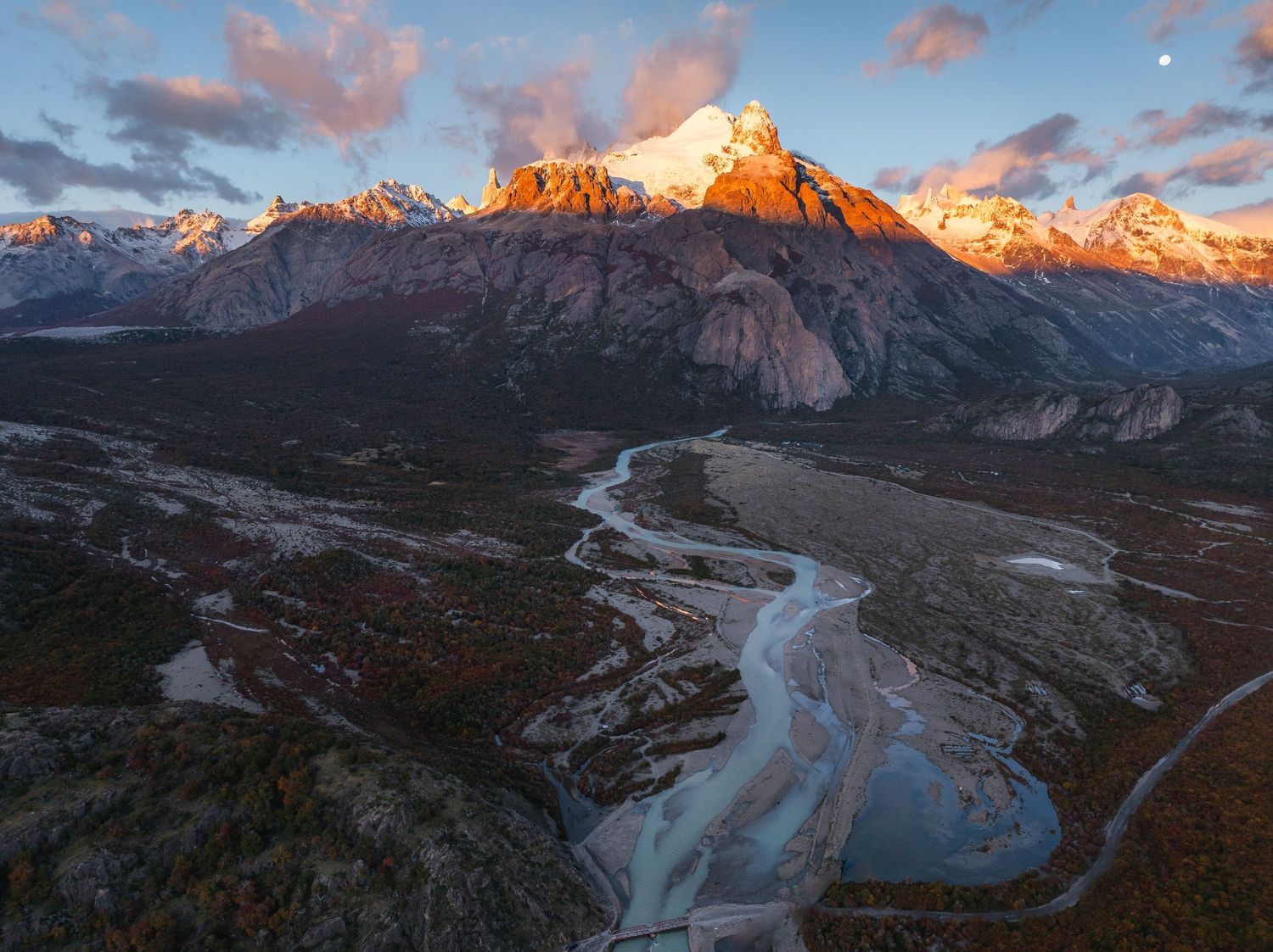 Aerial view of a winding glacial river at sunset with snow-capped mountain peaks glowing orange in Patagonia.