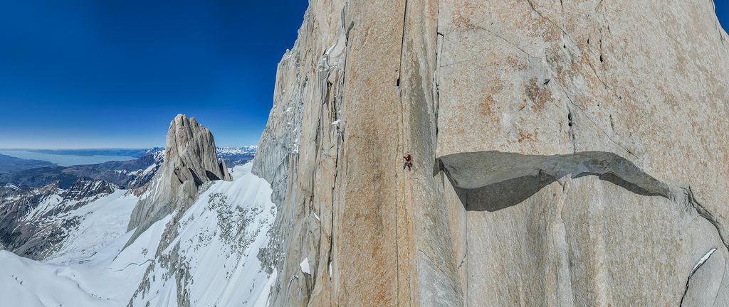 Aerial panoramic view of a climber on a sheer granite wall with snowy Patagonian landscape stretching below.