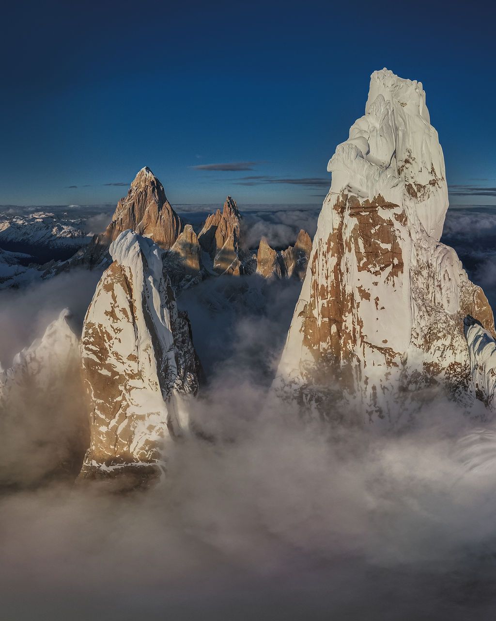 Massive ice-covered granite towers of Patagonia pierce through swirling clouds under a deep blue sky.